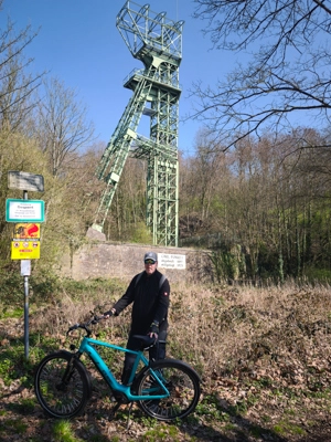Eine Person steht mit einem Fahrrad auf einem Waldweg. Hinter ihr steht ein großes grünes Stahlgerüst, das wie ein alter Förderturm aussieht. Bäume und trockenes Laub umgeben die Szene, links stehen mehrere Schilder am Weg.